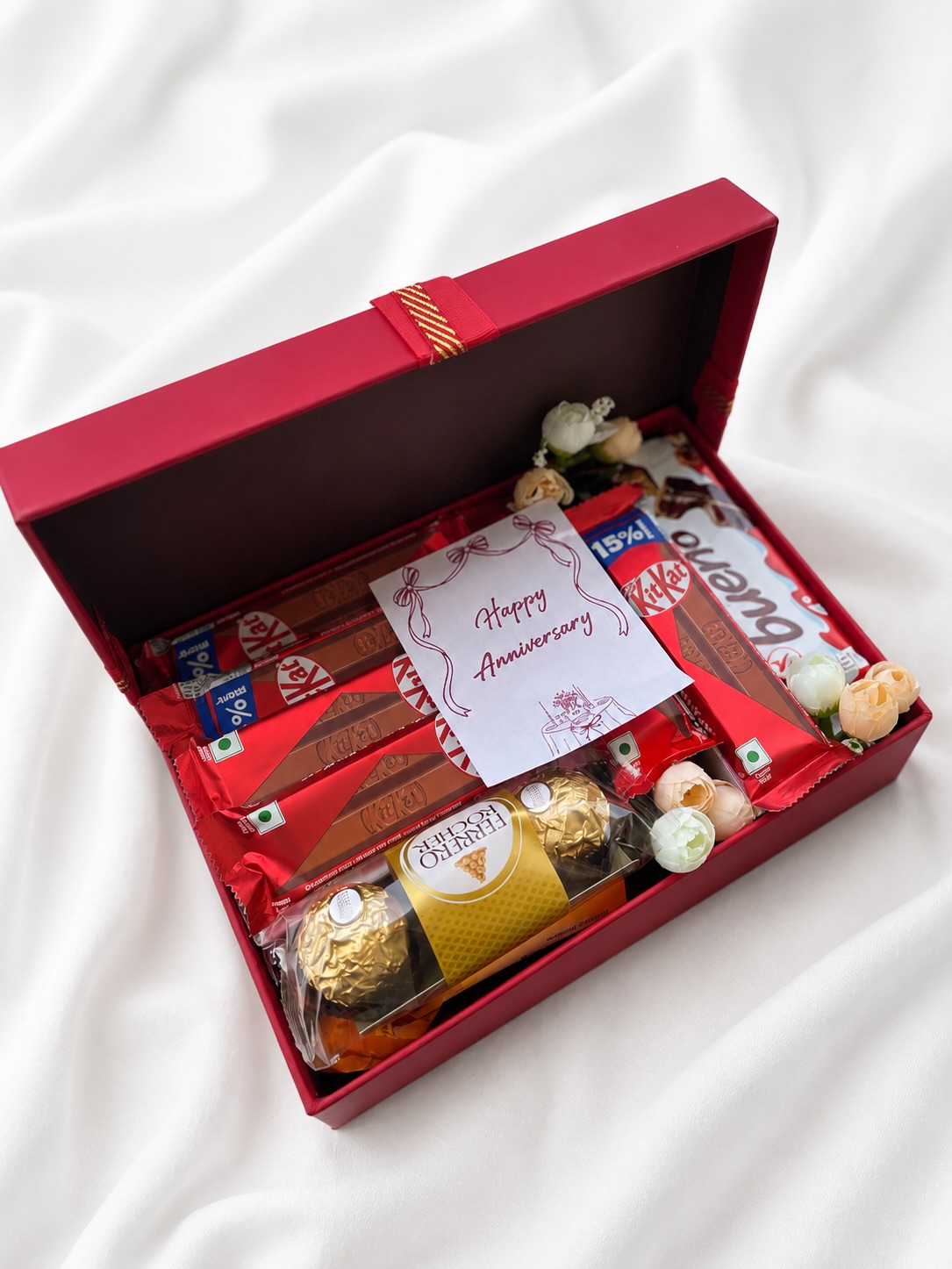 A red keepsake box layered with KitKat bars, Ferrero Rocher, Guylian chocolate, and cream florals, topped with a Happy Anniversary card.