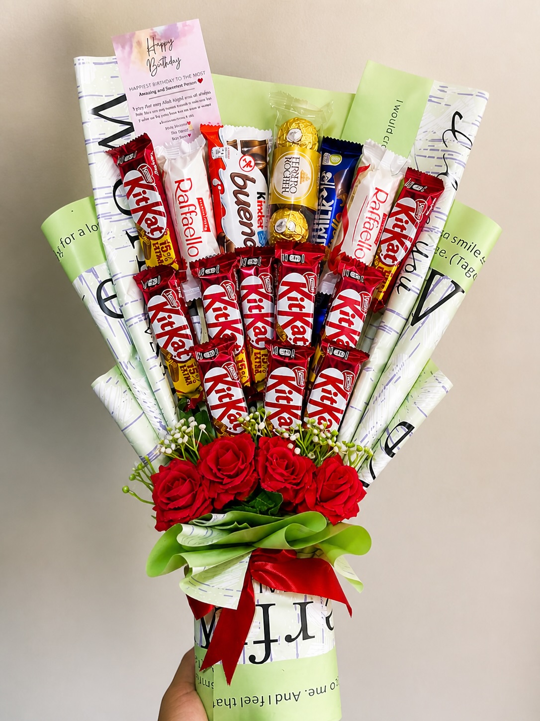 A tall chocolate bouquet of KitKat, Ferrero Rocher, Kinder Bueno, and Raffaello bars with red roses and a Happy Birthday message card.