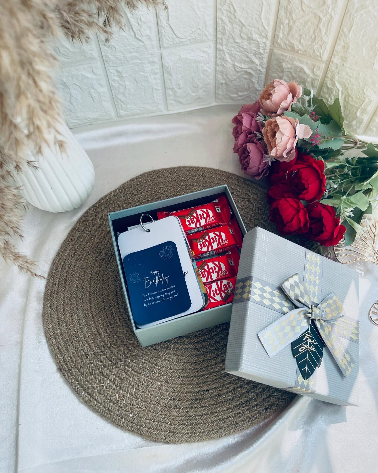 A sage gift box lined with KitKat bars and a handwritten-style birthday message card, wrapped alongside a diamond-ribboned box and fresh roses.