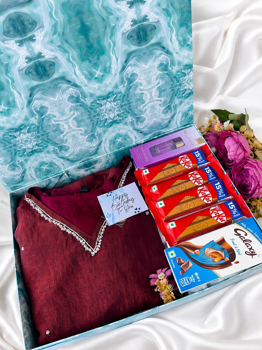 A generously stocked birthday box with an embellished kurti, perfume, and an assortment of KitKat and Galaxy chocolates.