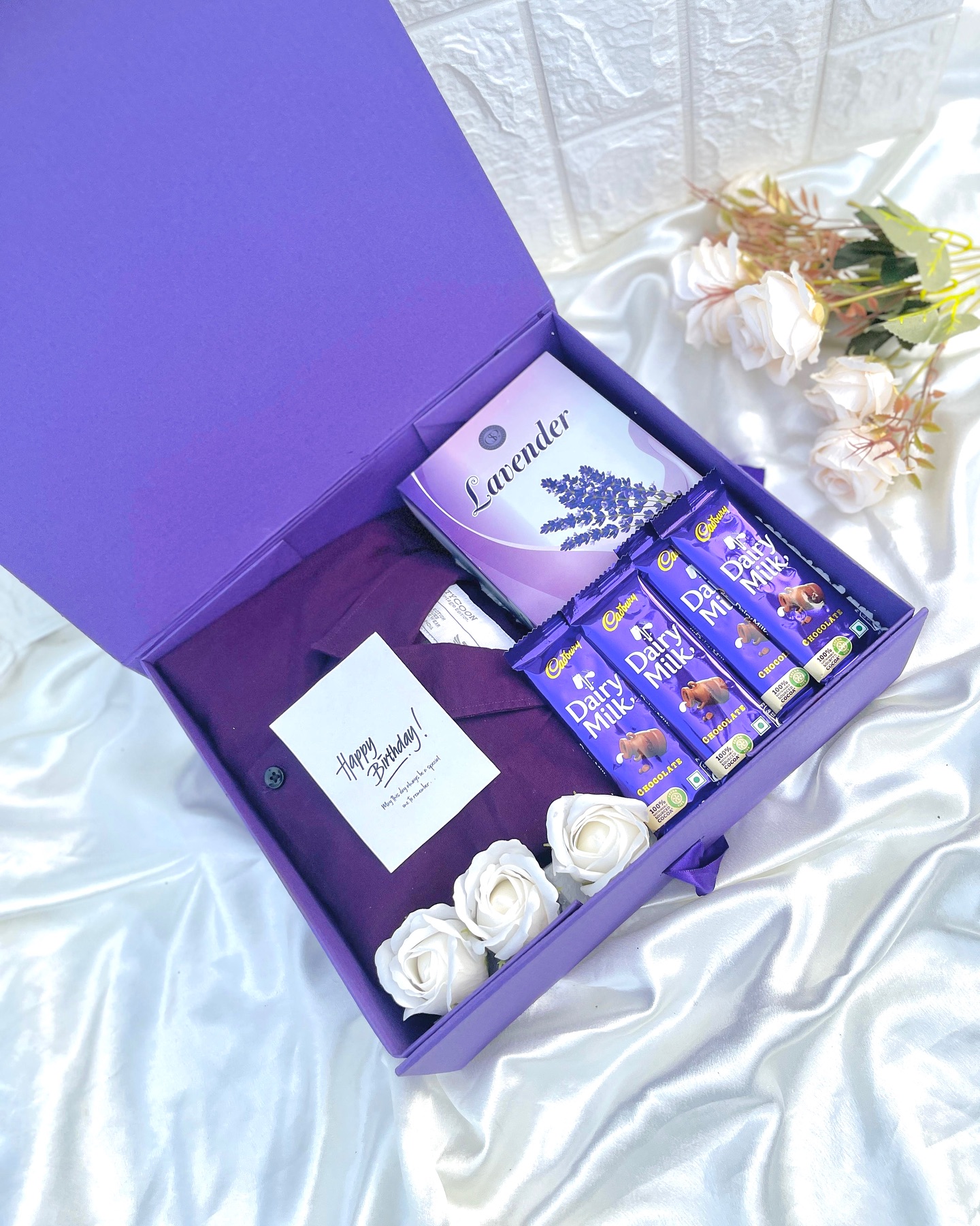 A rich plum gift box pairing a soft shirt with lavender, white roses, and Cadbury chocolates.