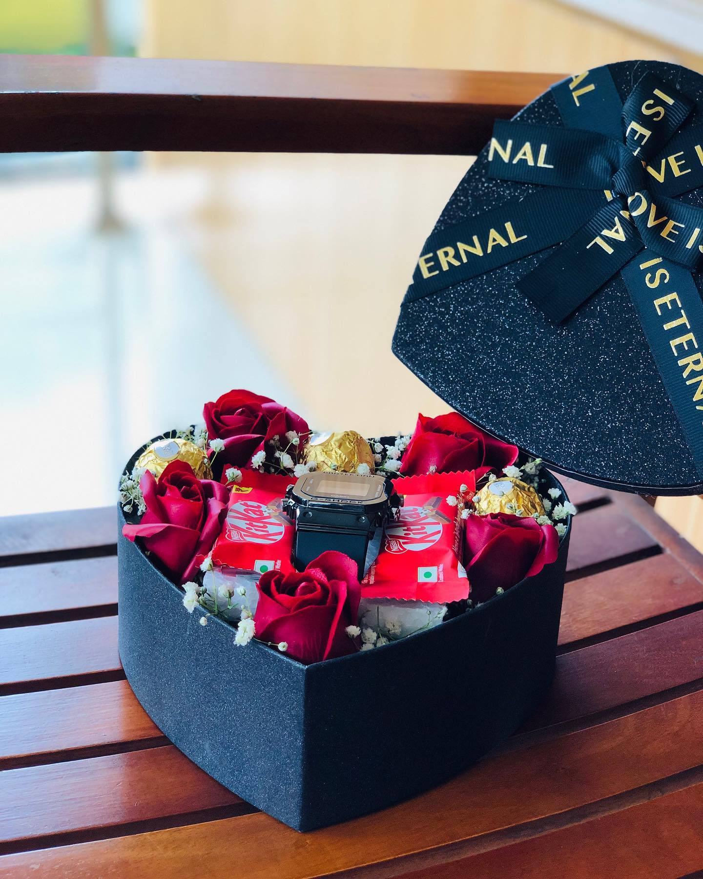 A glittering black heart-shaped box filled with red roses, Ferrero Rocher, KitKat, and a wristwatch.