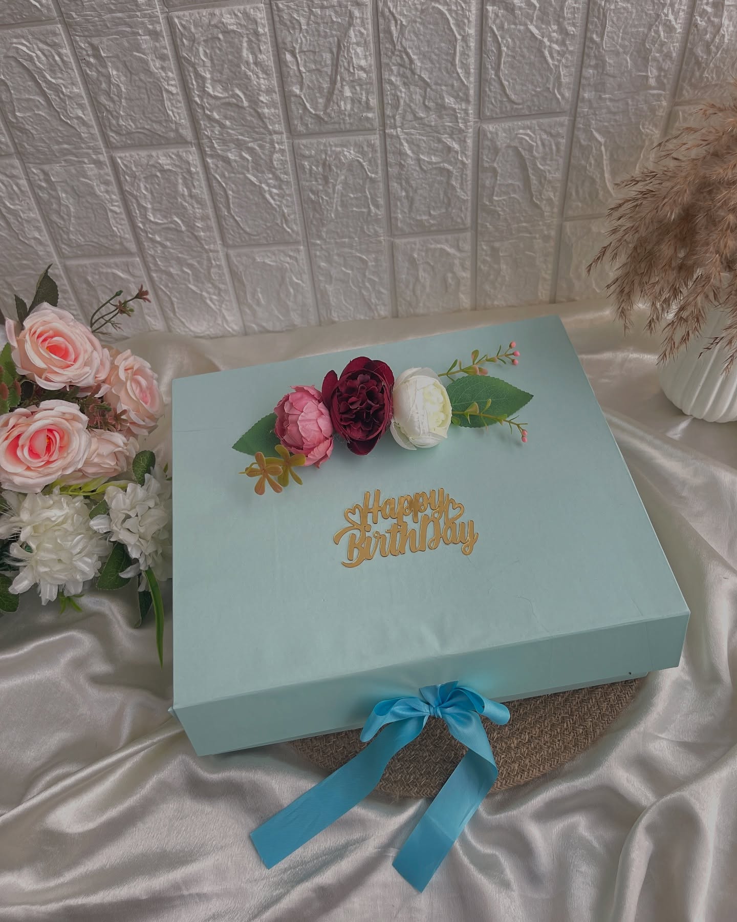 An elegant mint-blue gift box topped with silk peonies and a gold 'Happy Birthday' topper, tied with a satin ribbon.