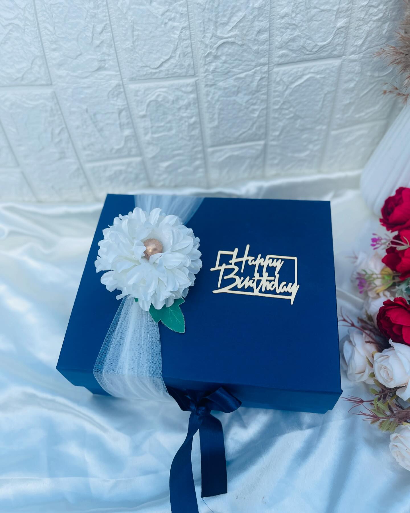 A deep navy keepsake box wrapped in tulle with a white blossom and a gold Happy Birthday charm, framed by fresh roses.
