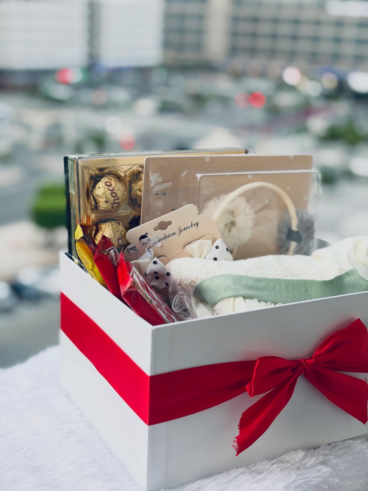 A white-and-red gift box filled with soft baby essentials, a delicate headband, and Ferrero Rocher chocolates.