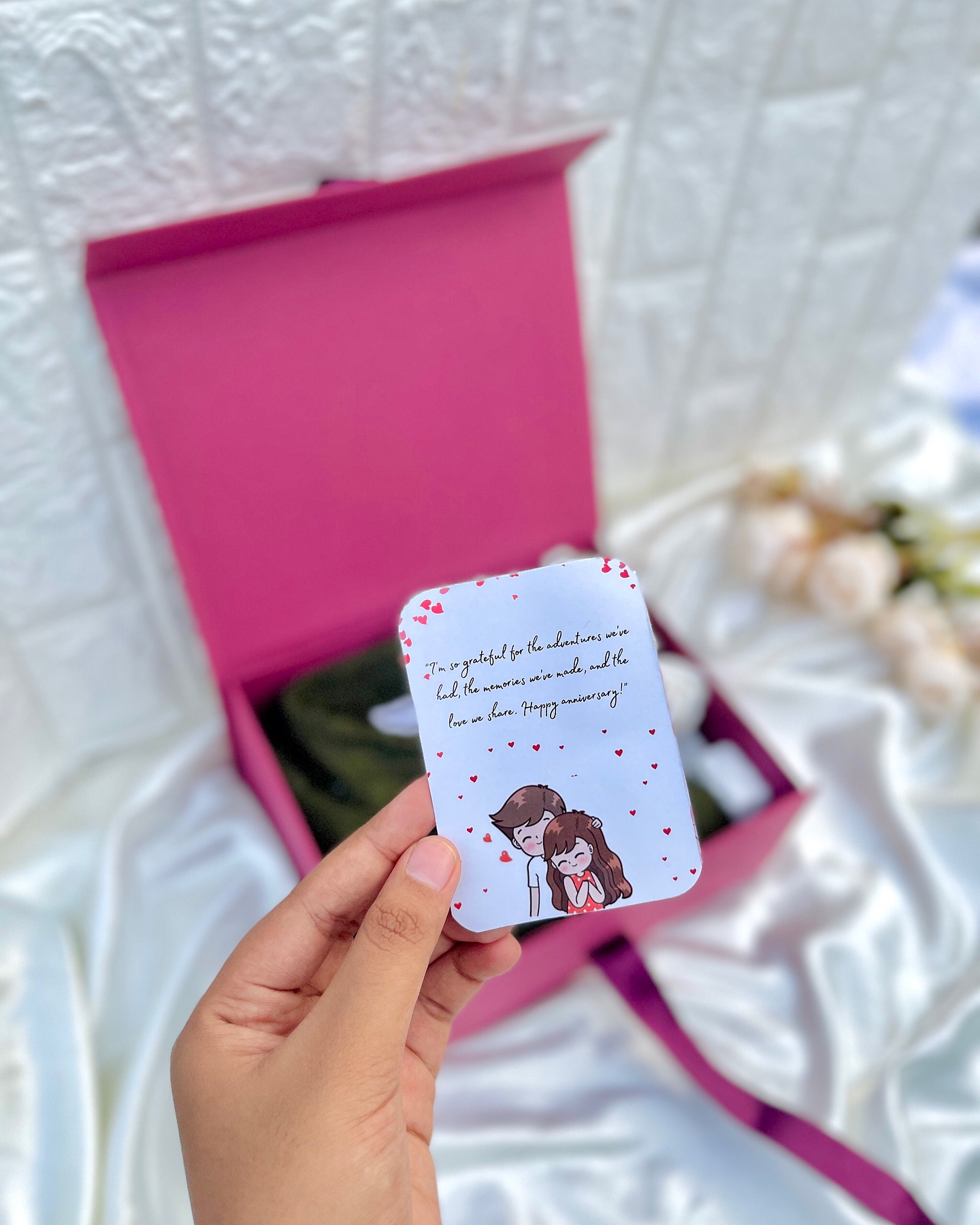 A pink keepsake box holding a soft olive cardigan, white silk roses, and a heartfelt anniversary message card.