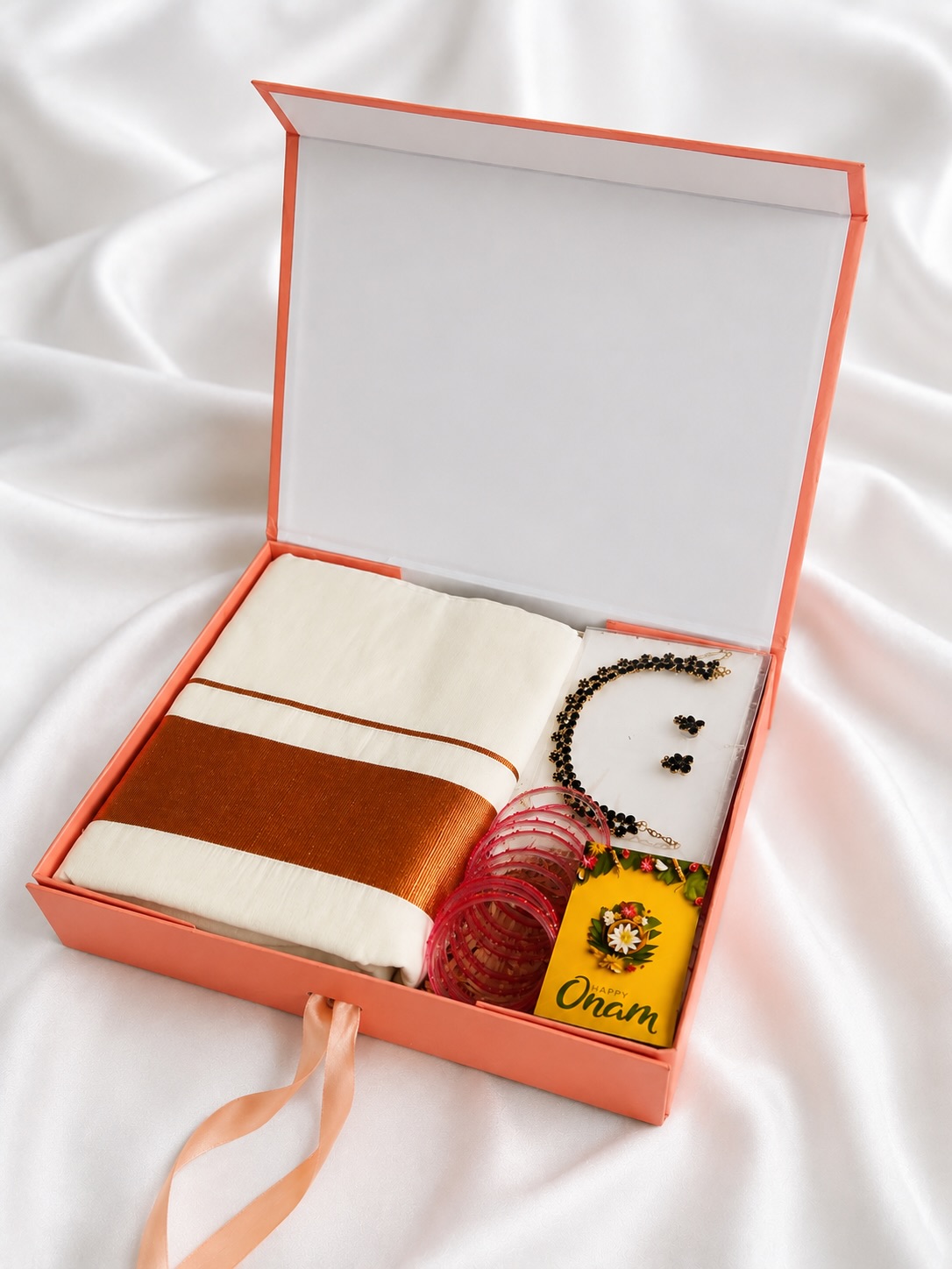 A festive Onam gift box with a traditional kasavu-bordered mundu, an elegant beaded jewelry set, and pink bangles.
