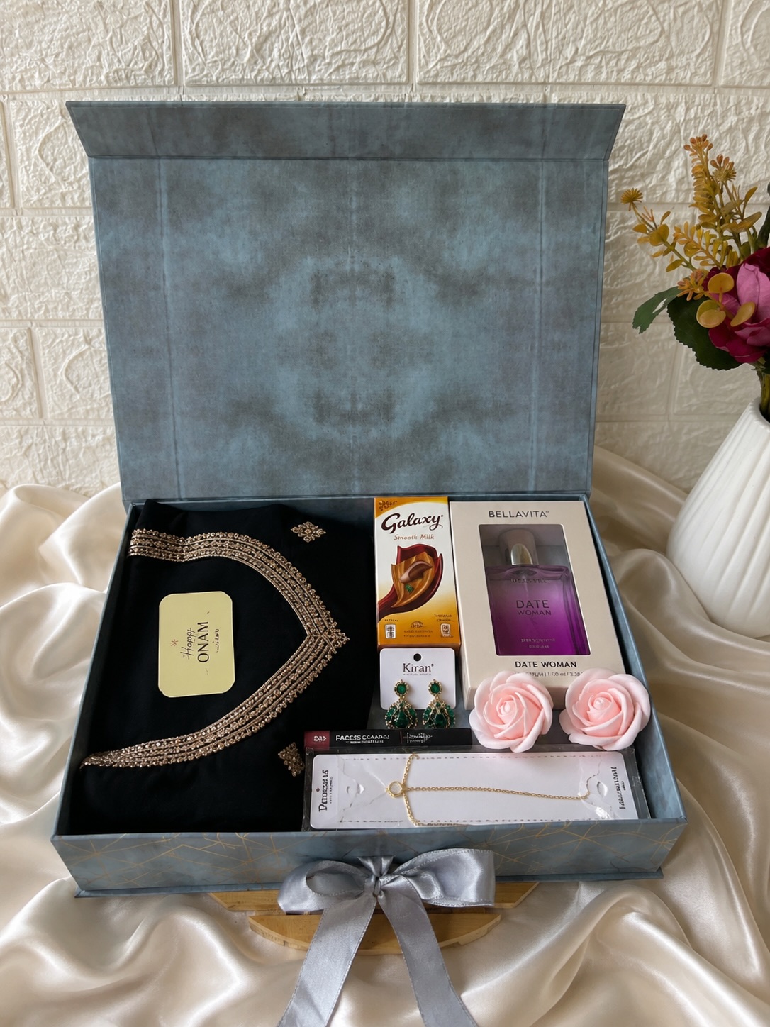 A festive grey gift box layering a black embroidered fabric piece with perfume, jewelry, and chocolates for Onam.