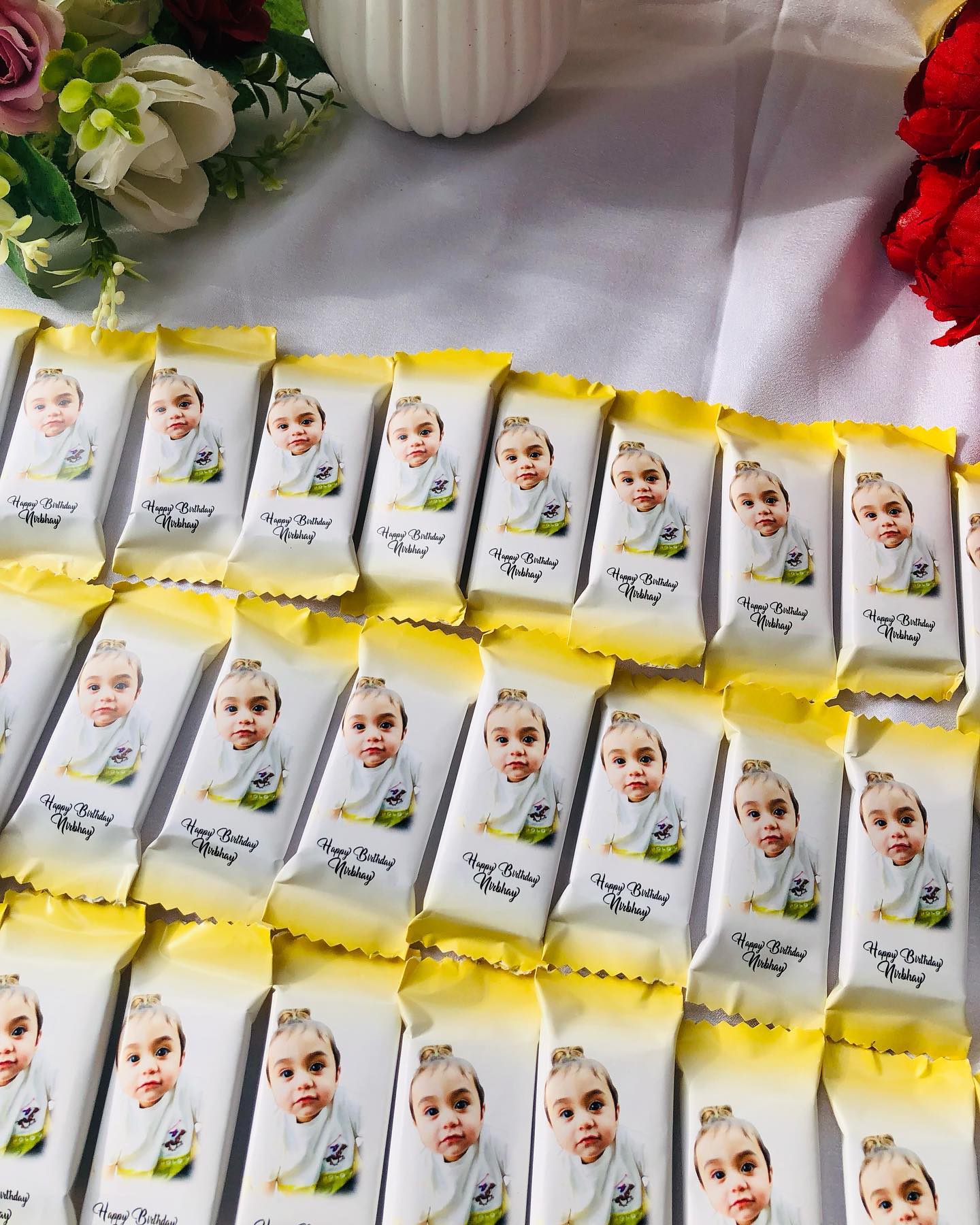 A tray of mini chocolate bars individually wrapped with a baby's photo and 'Happy Birthday' name print in cheerful yellow packaging.