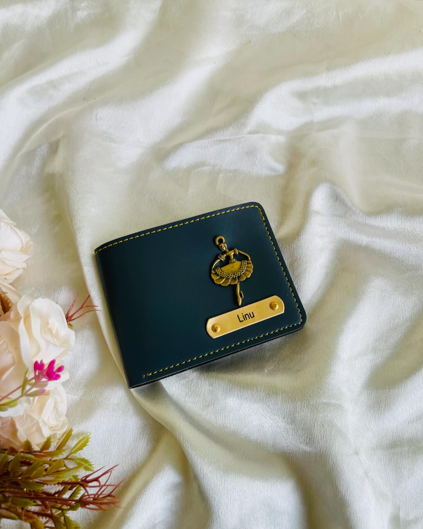 A teal leather bifold wallet finished with a gold ballerina charm and an engraved name plate.