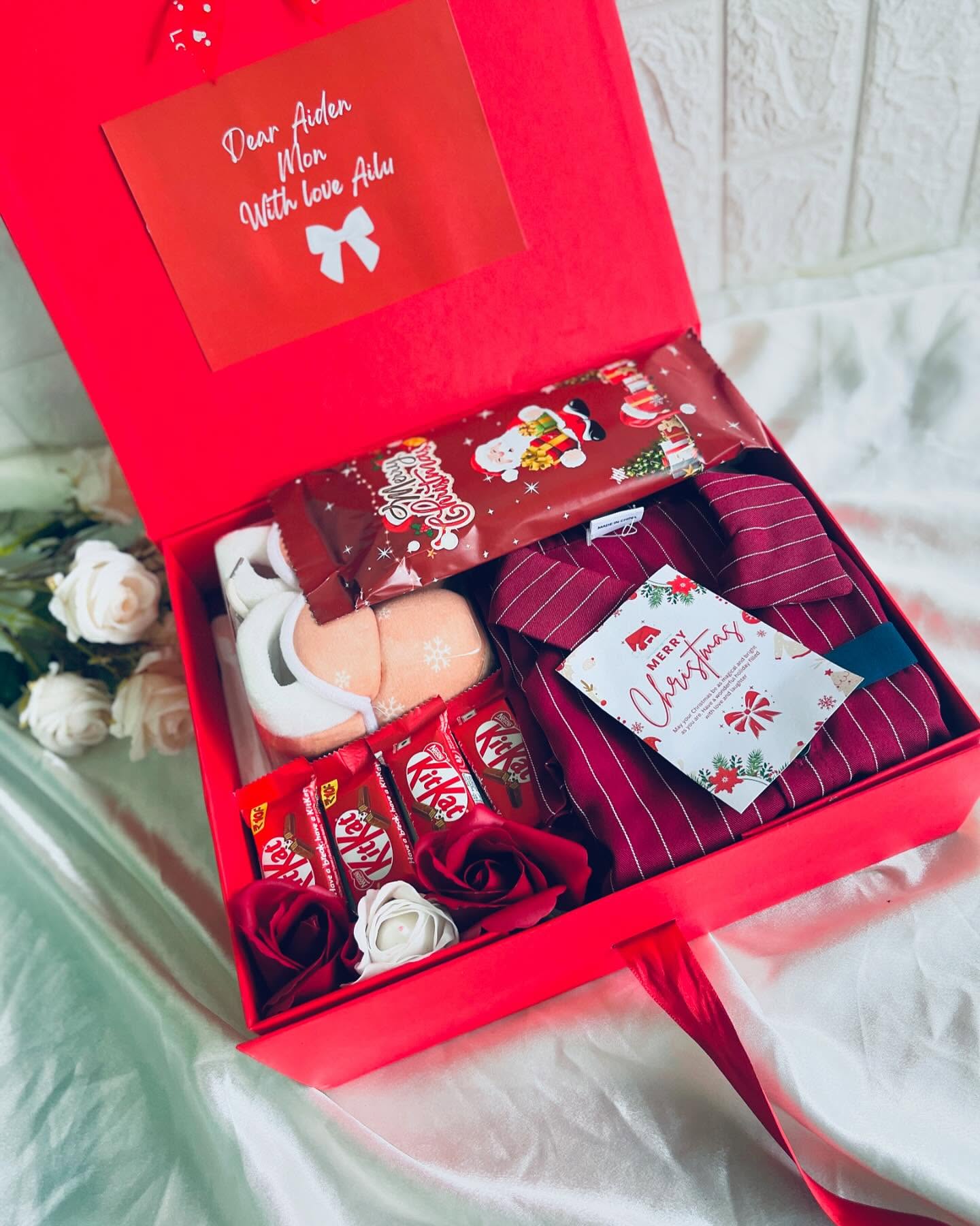 A red Christmas gift box with a name-dedicated card, baby booties, a striped tie and pajama set, KitKat bars and a 'Merry Christmas' note.