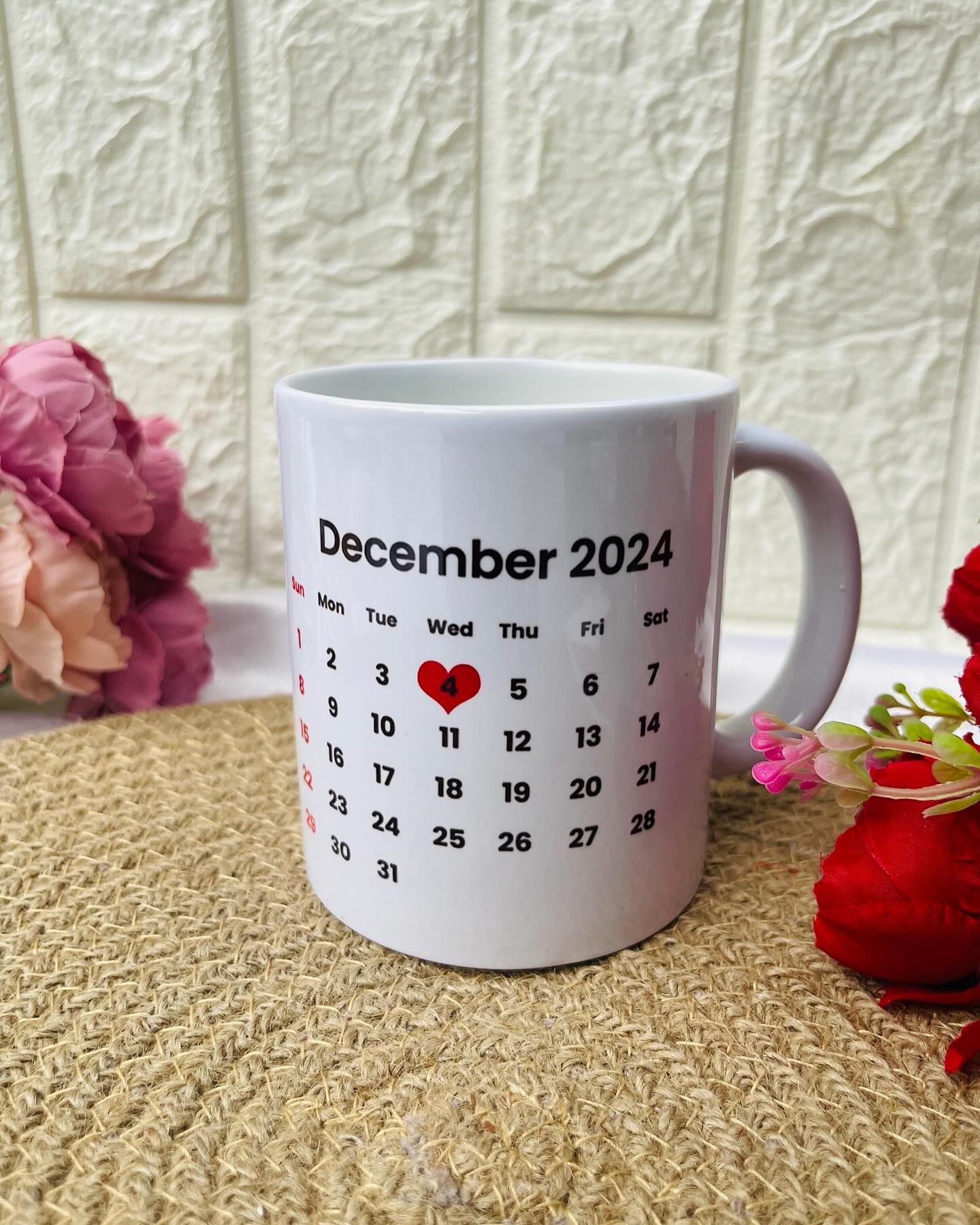 A white ceramic mug printed with a custom monthly calendar, marking a special date with a red heart.