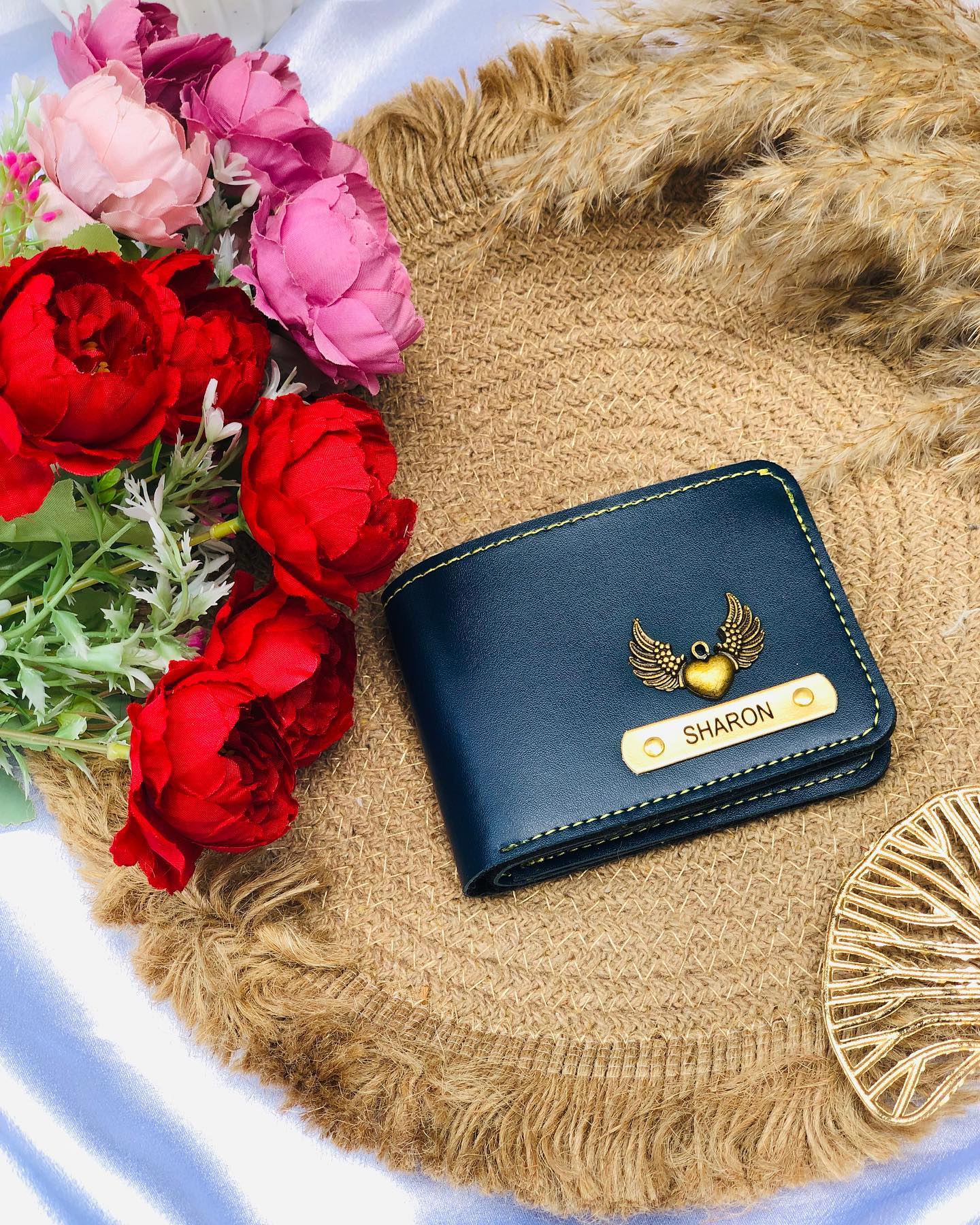 A navy leather wallet personalized with the name "Sharon" and a heart-wing charm, styled with fresh roses and peonies.