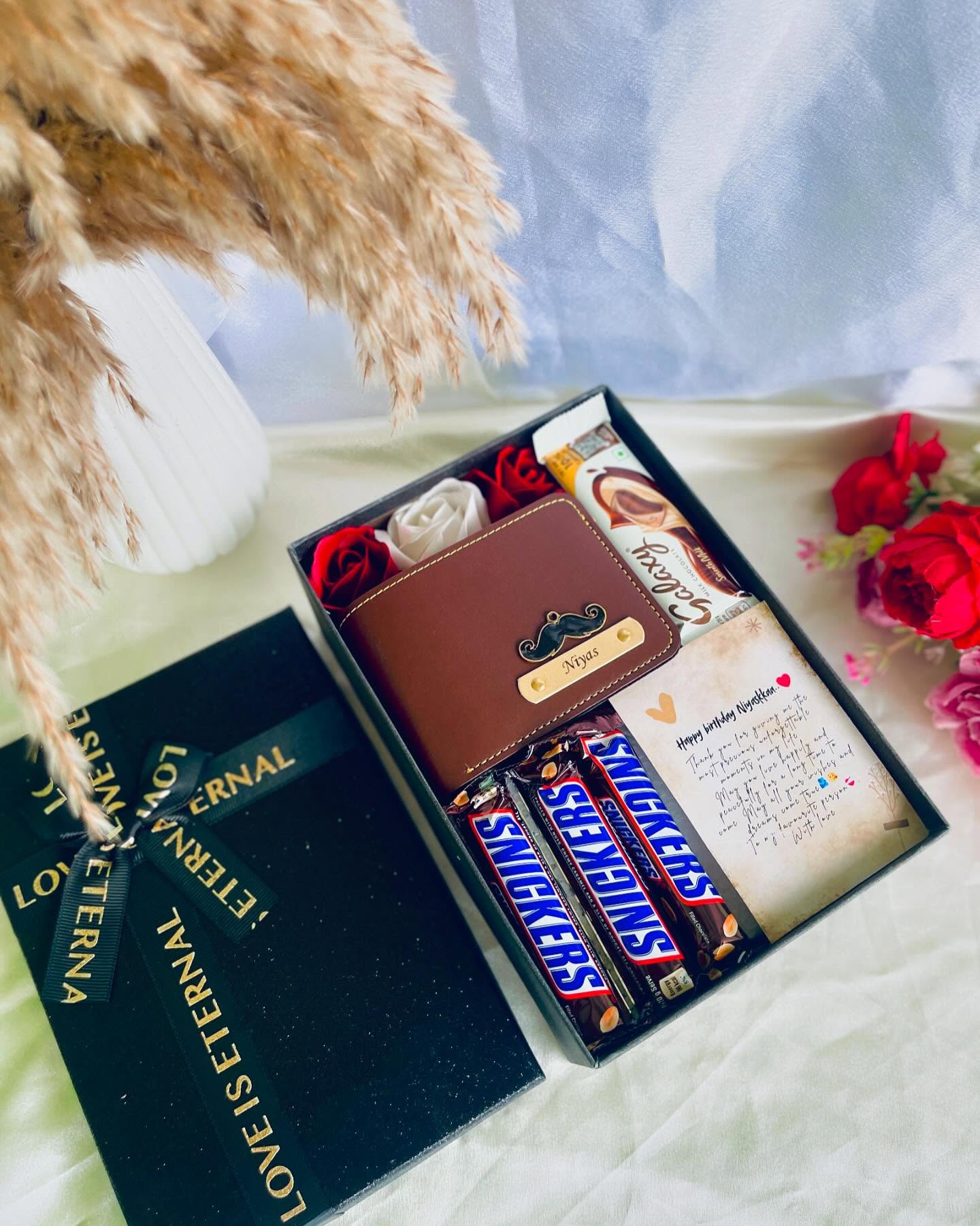 A black "Love is Eternal" gift box with a name-embossed leather wallet, roses, chocolates, and a handwritten birthday note.