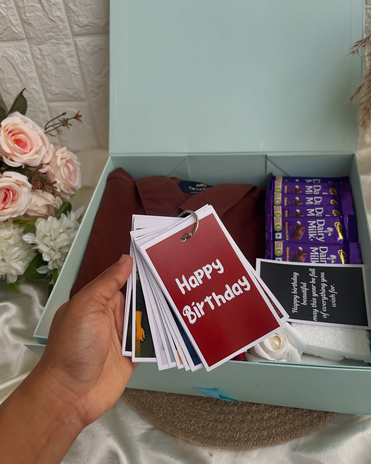 A mint gift box with a folded shirt, Cadbury chocolates, and a ring-bound photo flip book for a birthday.