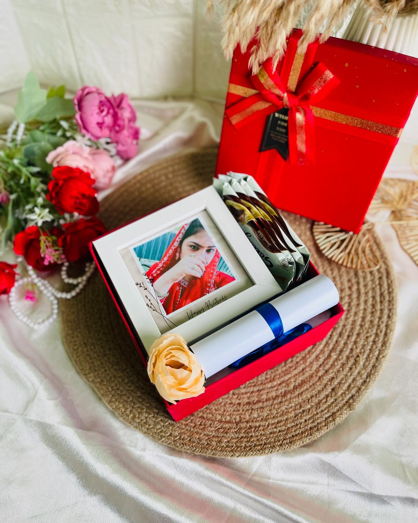 A red keepsake box with a personalized photo frame, snack bars, a rolled certificate, and fresh flowers.