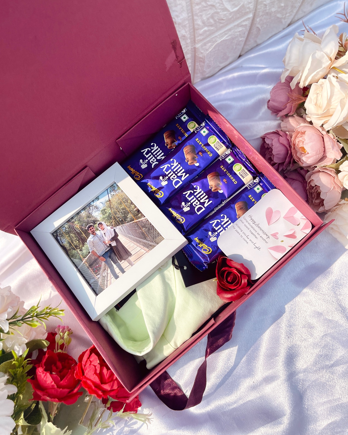 An anniversary gift box with a personalized couple photo frame, Dairy Milk chocolates, a soft shawl, and roses.