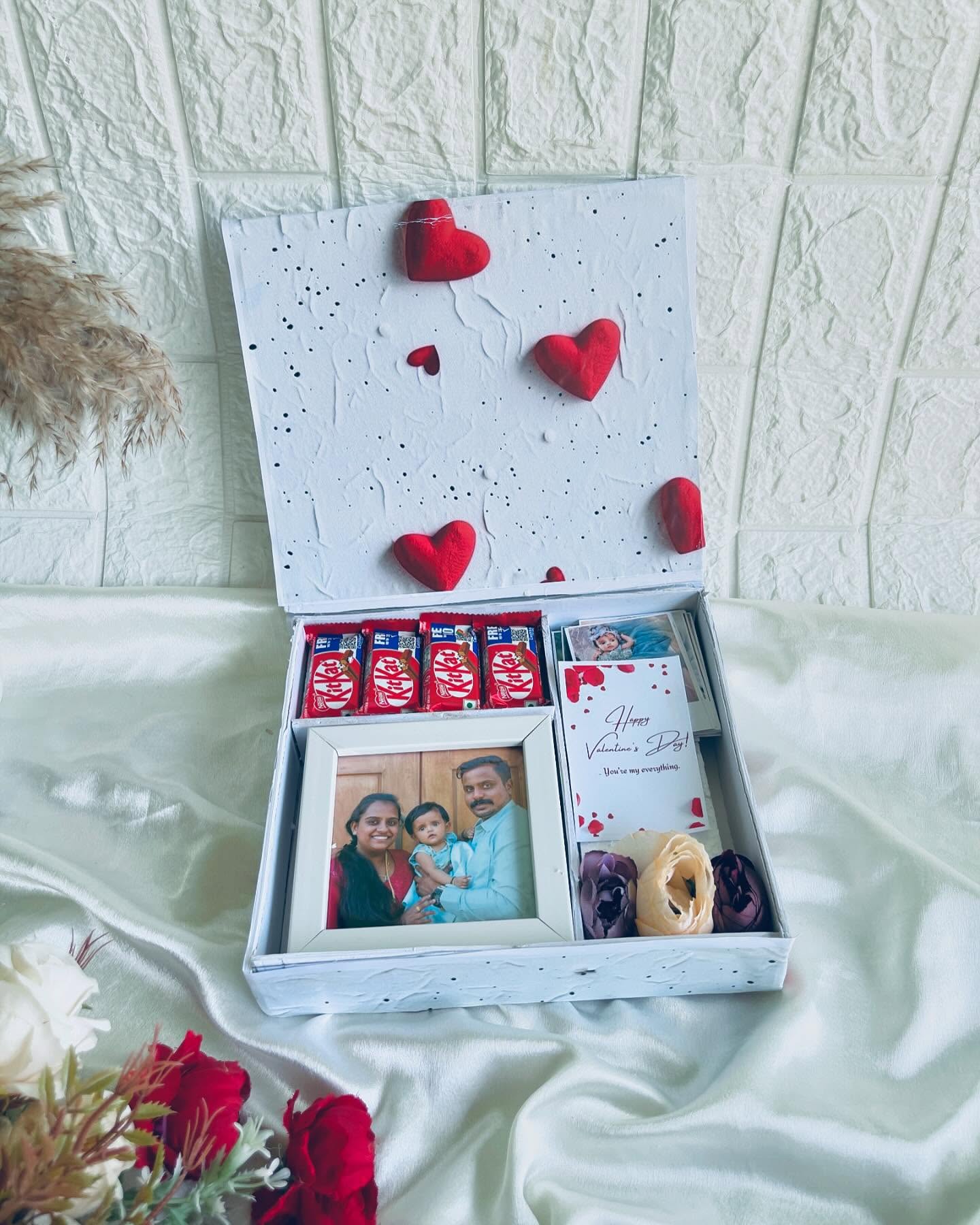 A heart-themed gift box featuring a personalized photo frame, KitKat chocolates, dried flowers, and a handwritten note card.