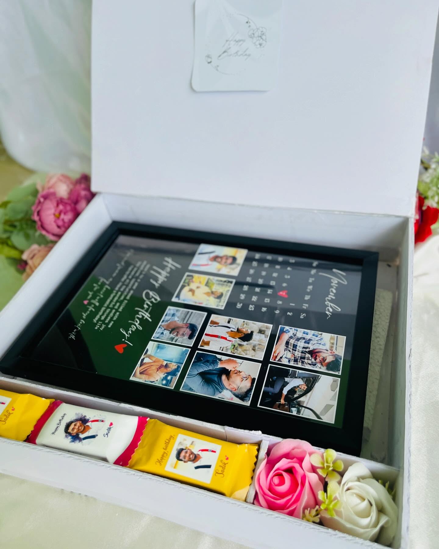 A birthday box holding a personalized photo-collage calendar frame alongside custom-wrapped chocolates and roses.