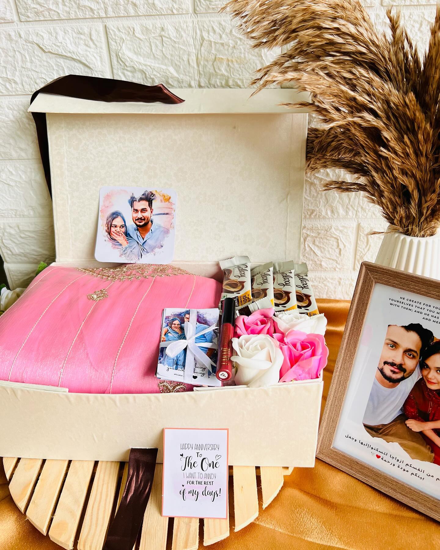 An open keepsake box pairing an embroidered pink saree with Galaxy chocolate bars, fresh roses, a lip tint, and a personalized couple photo frame.