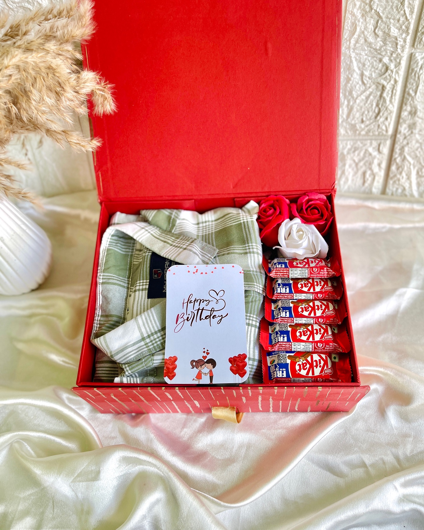 A red gift box filled with a checked pajama set, soap roses, a stack of KitKat bars, and a 'Happy Birthday' card.