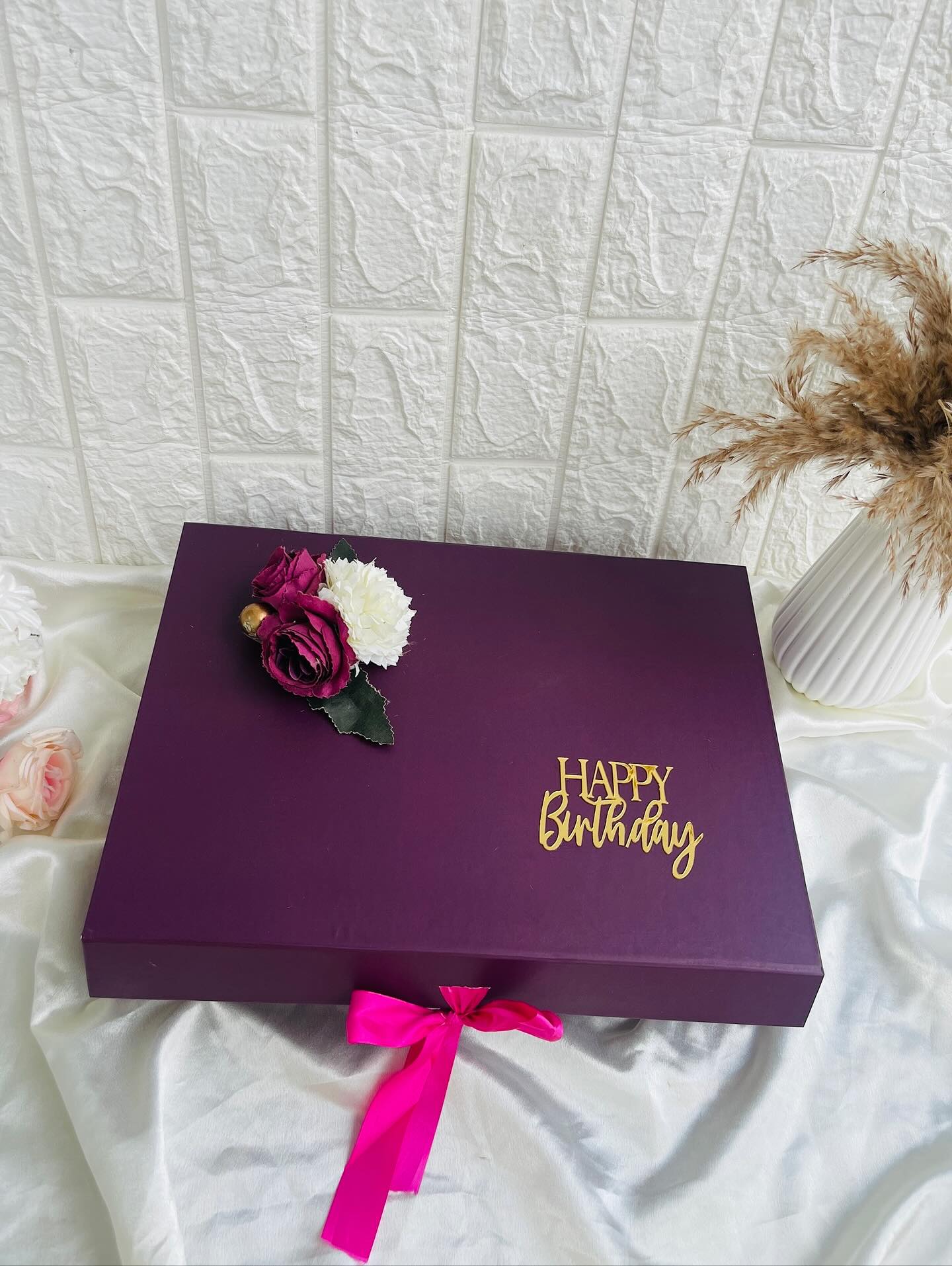 A deep plum gift box topped with maroon roses and a cream bloom, finished with a gold "Happy Birthday" topper and a hot pink ribbon.