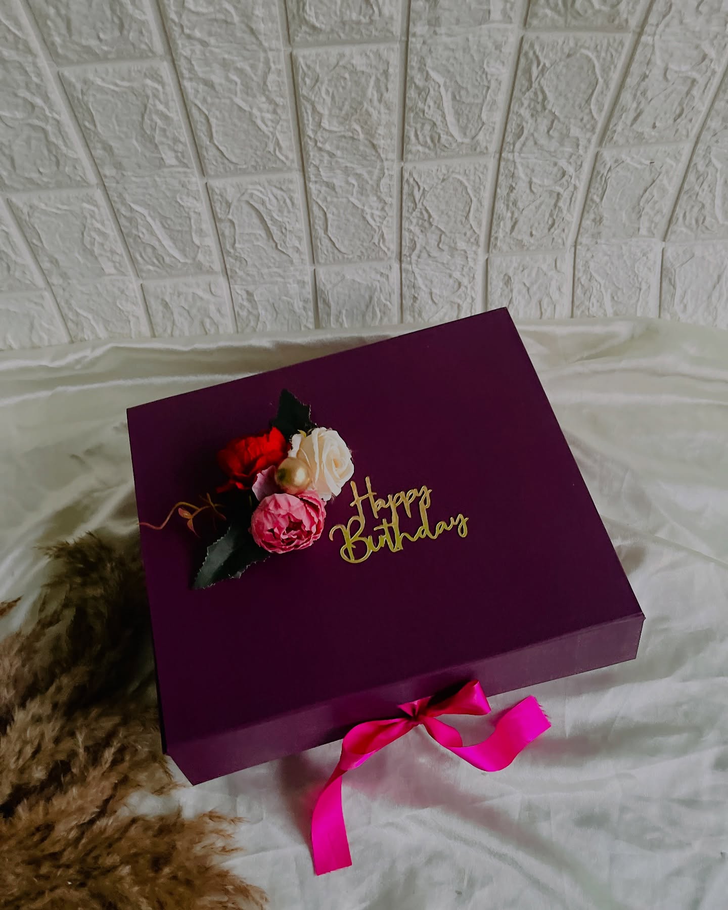 A rich plum gift box with a rose and peony accent and gold "Happy Birthday" script, tied with a pink satin bow.