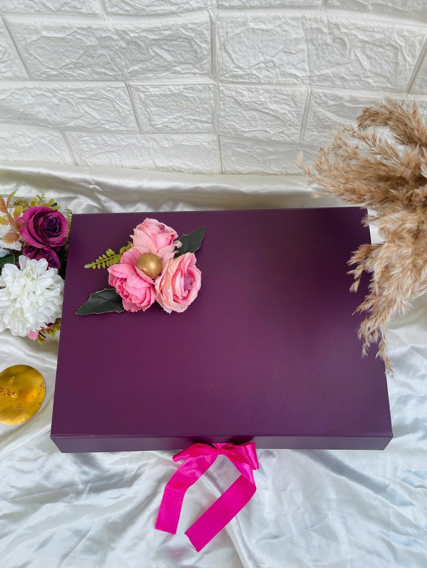 A rich plum-purple gift box adorned with a pink rose cluster and a hot-pink satin ribbon bow.