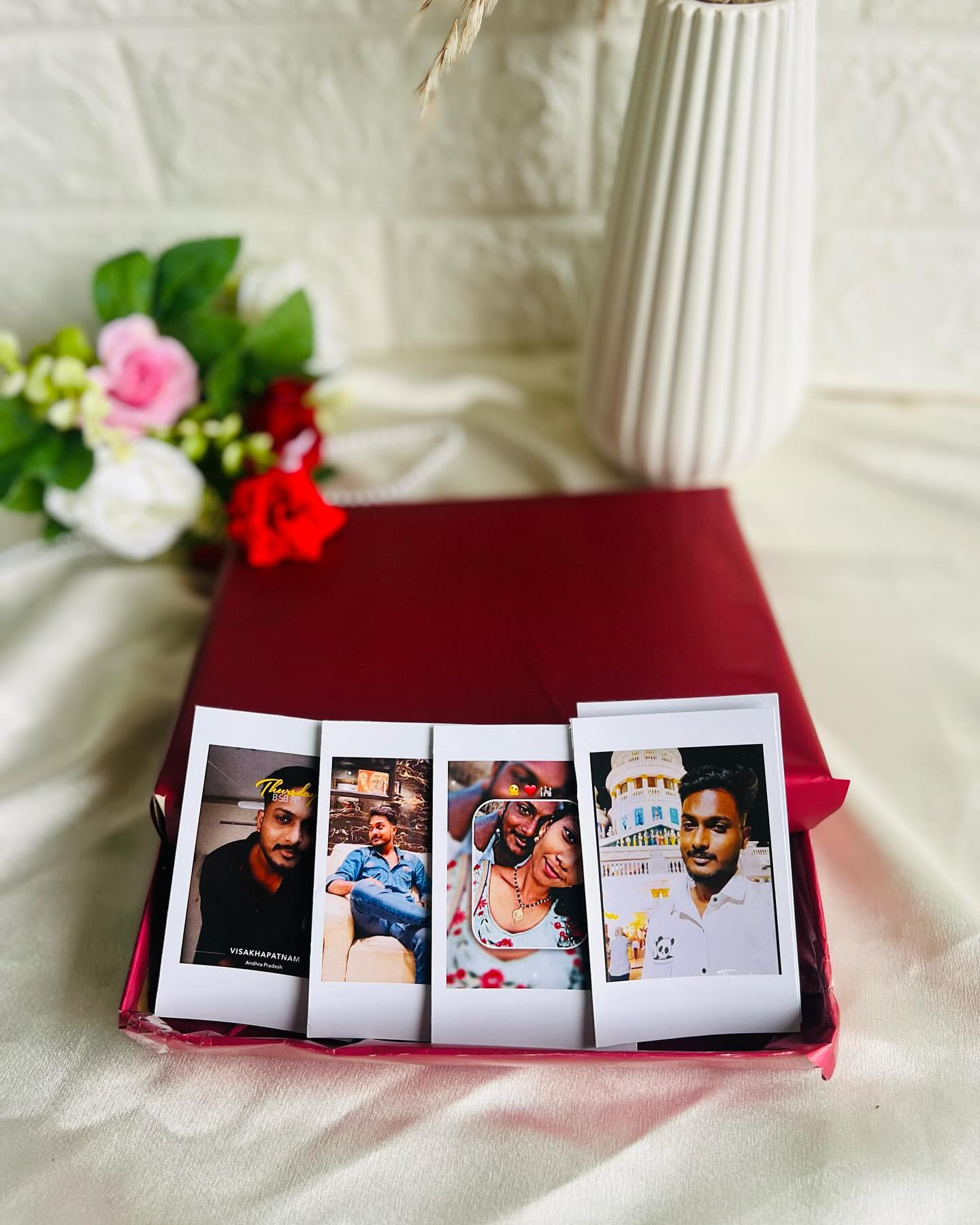 A wine-red gift box displaying a fanned-out set of polaroid-style personal photo prints alongside fresh florals.