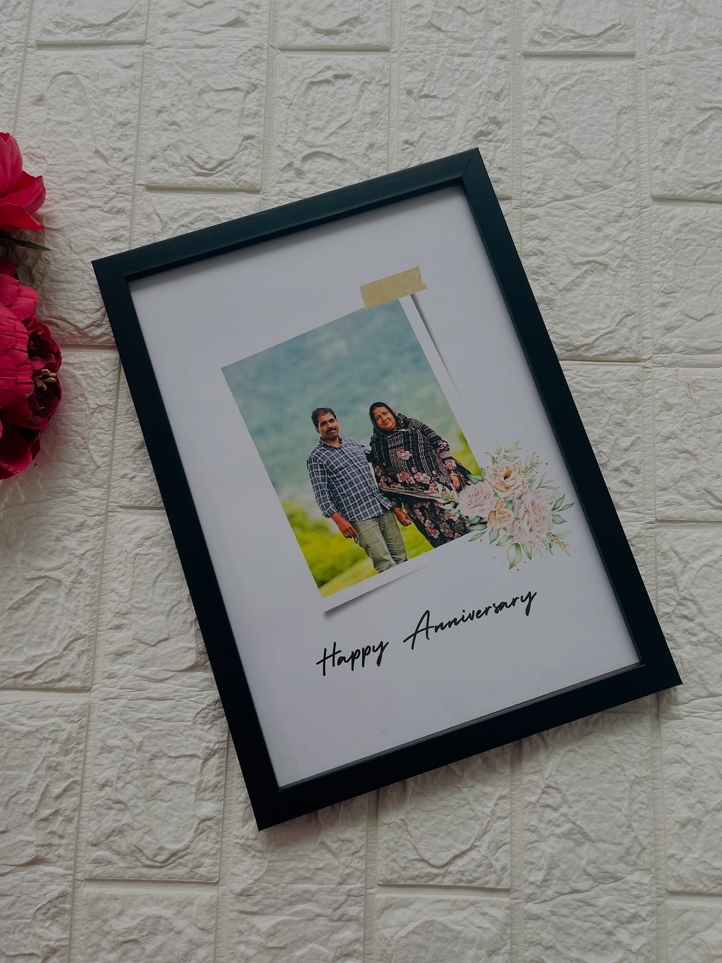 A black frame with a taped polaroid-style couple photo, florals, and a Happy Anniversary note.