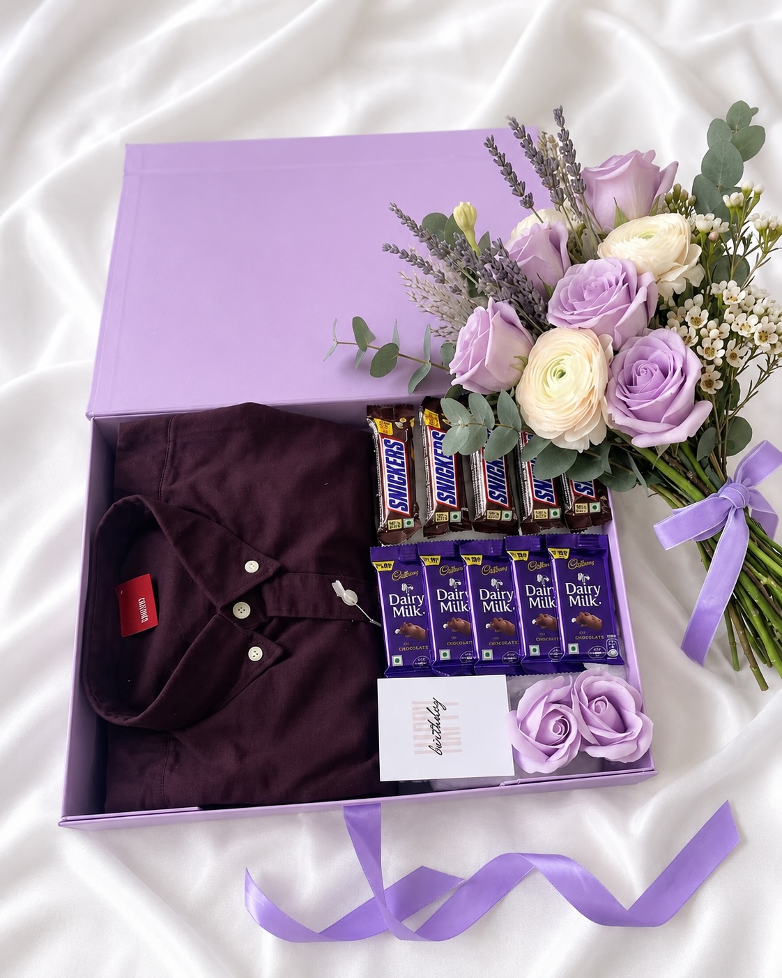 A lilac birthday box with a maroon polo shirt, Snickers and Dairy Milk bars, and a lavender-toned flower bouquet.