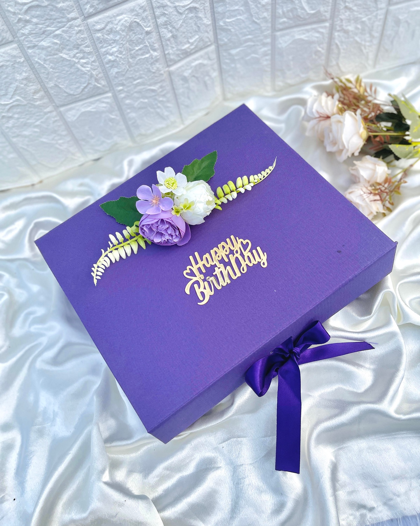 A rich purple gift box topped with a lasercut gold 'Happy Birthday' script and a delicate floral arrangement.