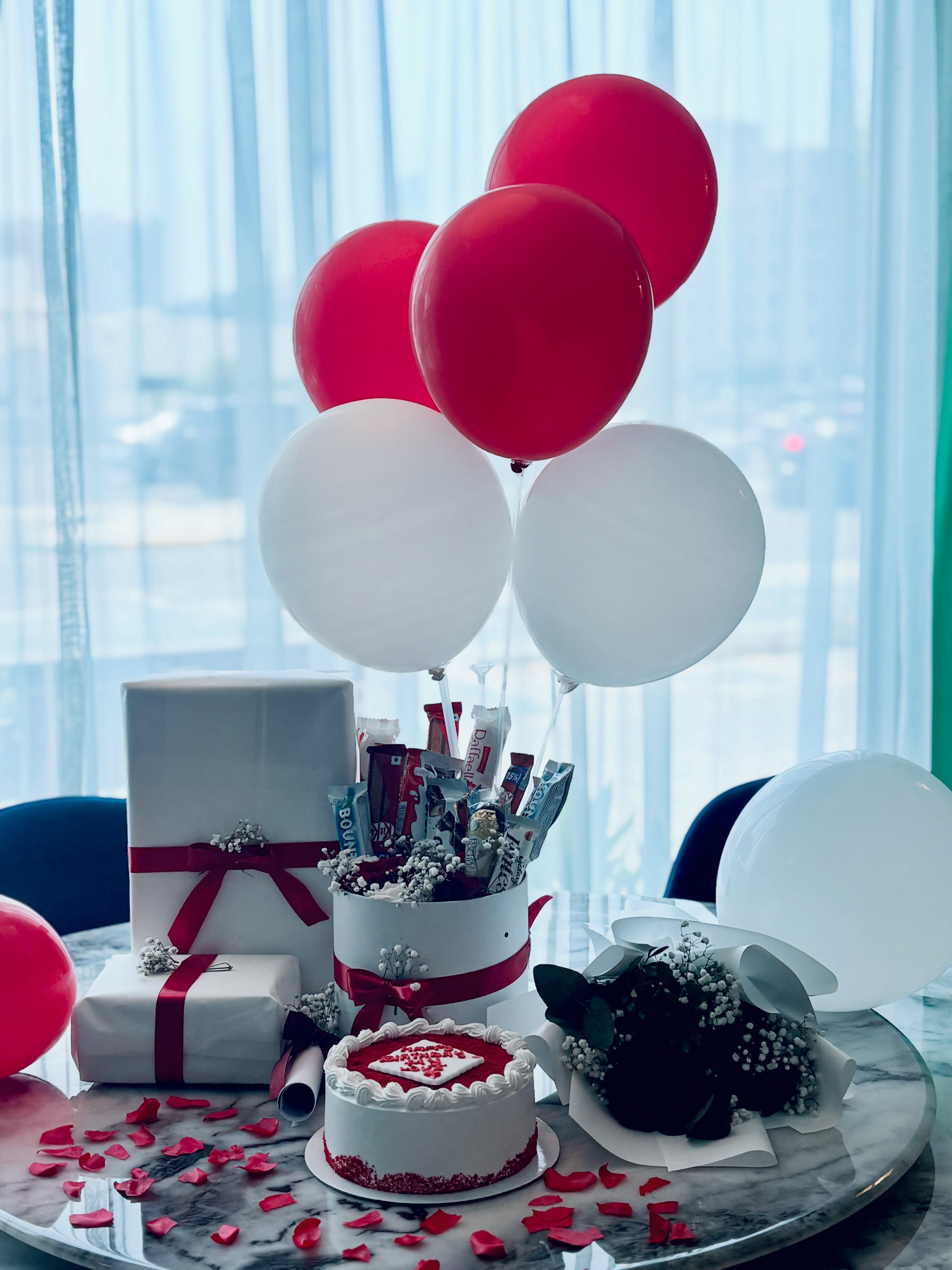 A full birthday surprise package with red and white balloons, wrapped gifts, a red velvet cake, a chocolate bouquet and fresh flowers.