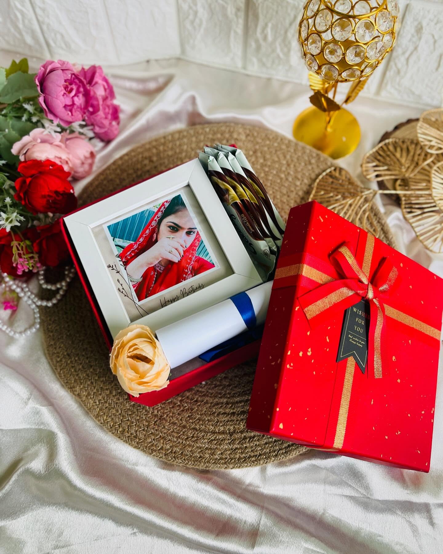 A red keepsake box with a personalized photo frame, chocolate bars, a rolled birthday scroll, and dried rose.