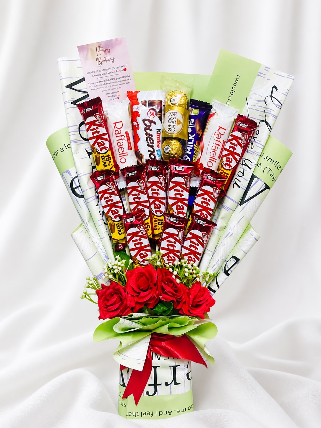 A tall chocolate bouquet of KitKat, Ferrero Rocher, and Kinder bars arranged with red roses and a birthday card.