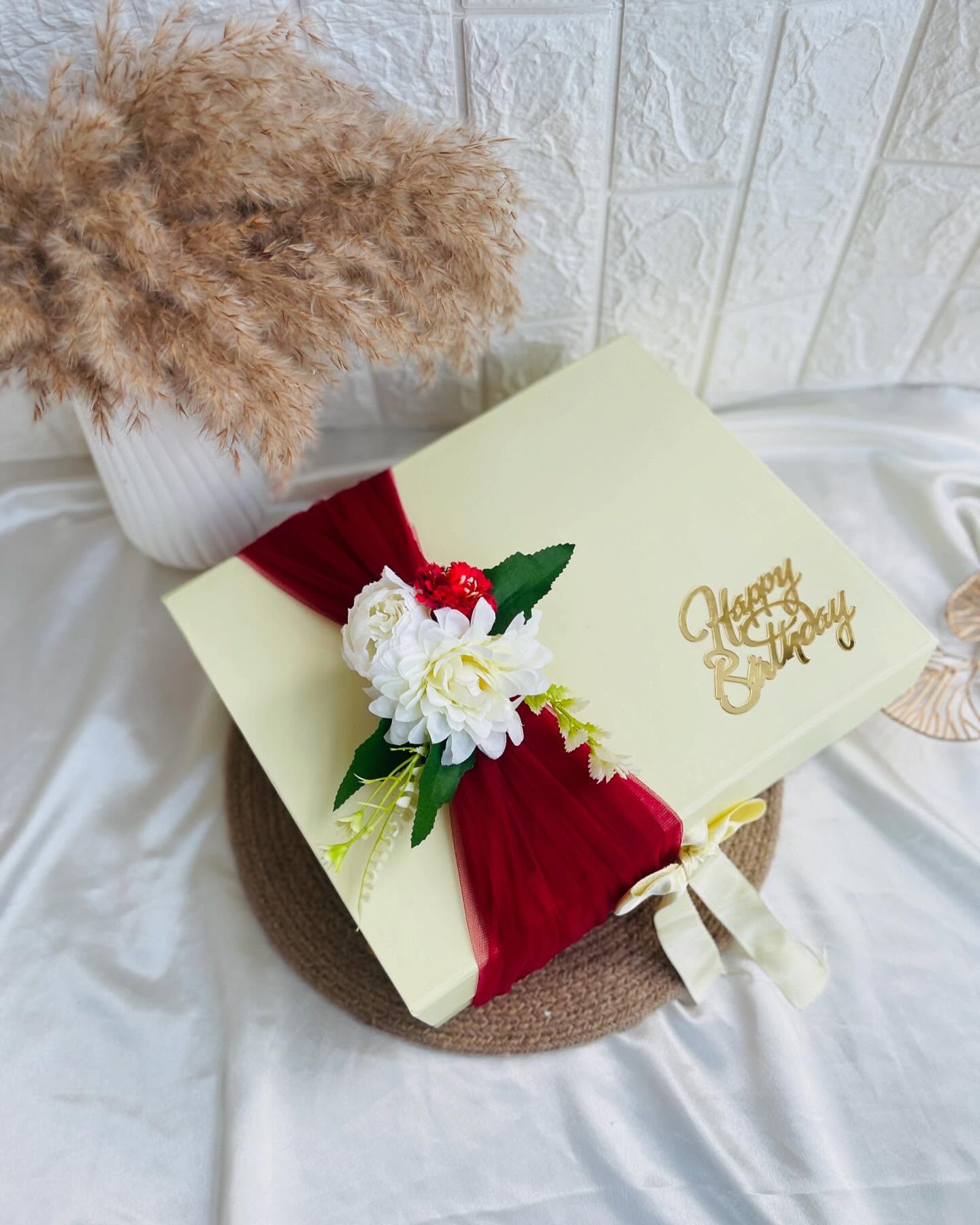 A cream gift box wrapped with a dramatic red tulle bow, white roses, and a gold 'Happy Birthday' topper.