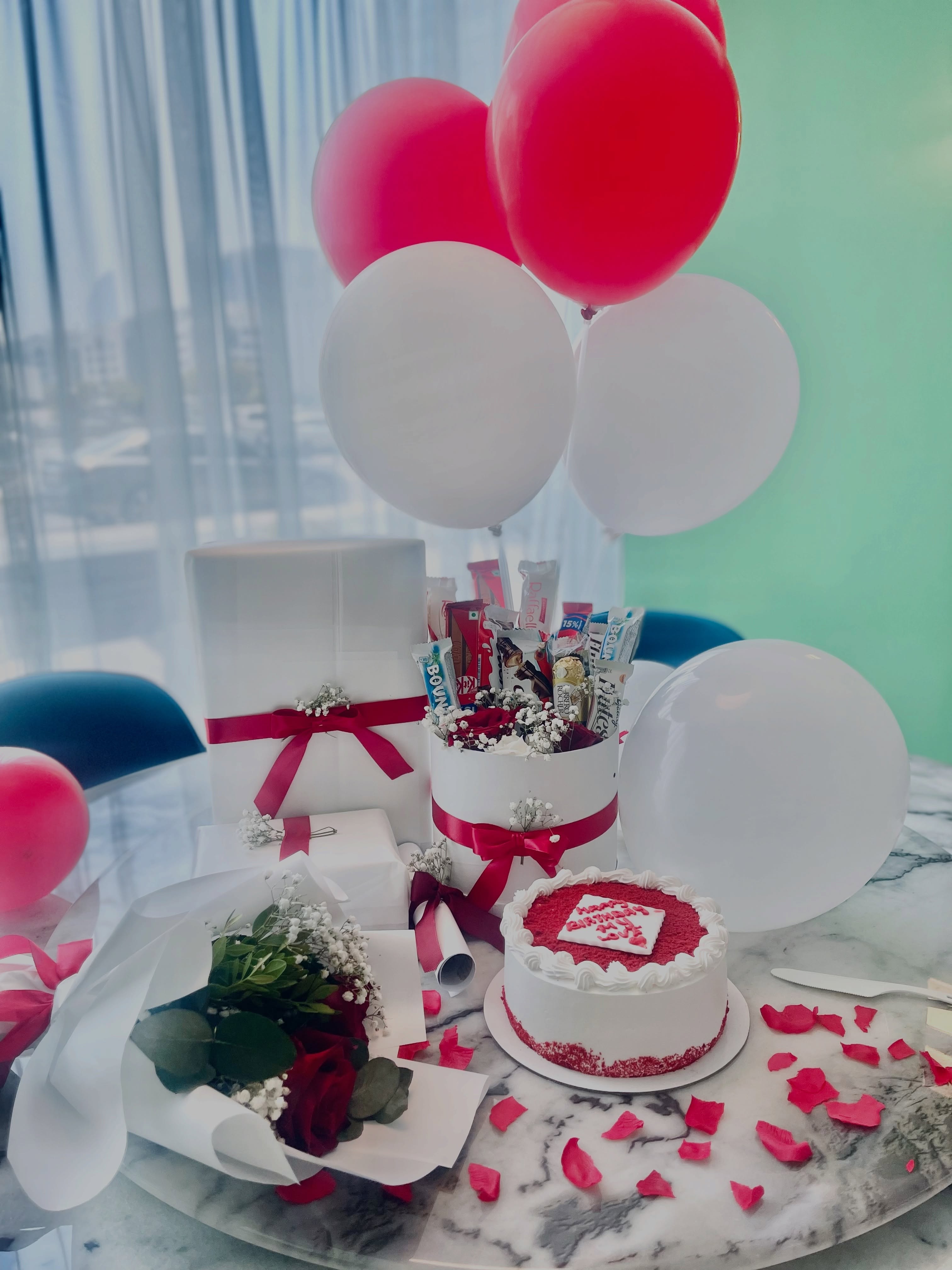 A full birthday celebration set with a red velvet cake, chocolate bouquet, roses, balloons, and gift boxes.