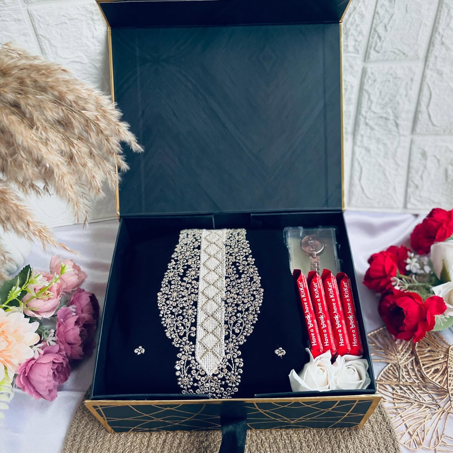 A luxurious black and gold gift box featuring a heavily embellished pearl-beaded stole, a wristwatch, earrings, and KitKats.
