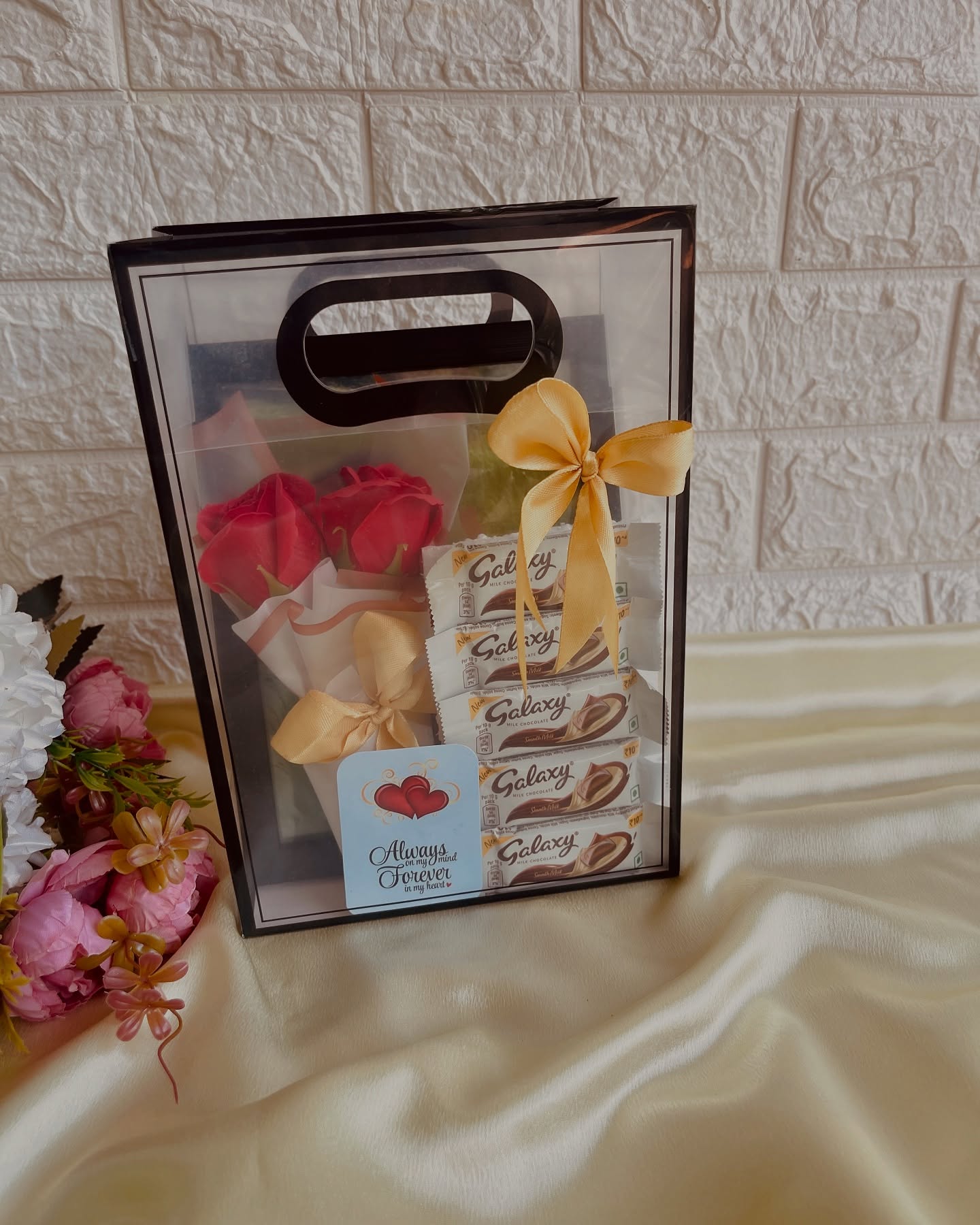 A clear gift bag holding red roses and Galaxy chocolate bars, tied with a gold satin bow and an "Always in my heart" message card.