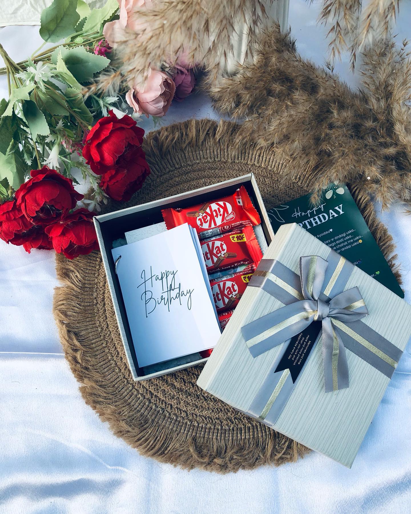 A jute-tray gift set with a KitKat-filled box, handwritten birthday card, and a ribboned gift box, styled with red roses and peonies.