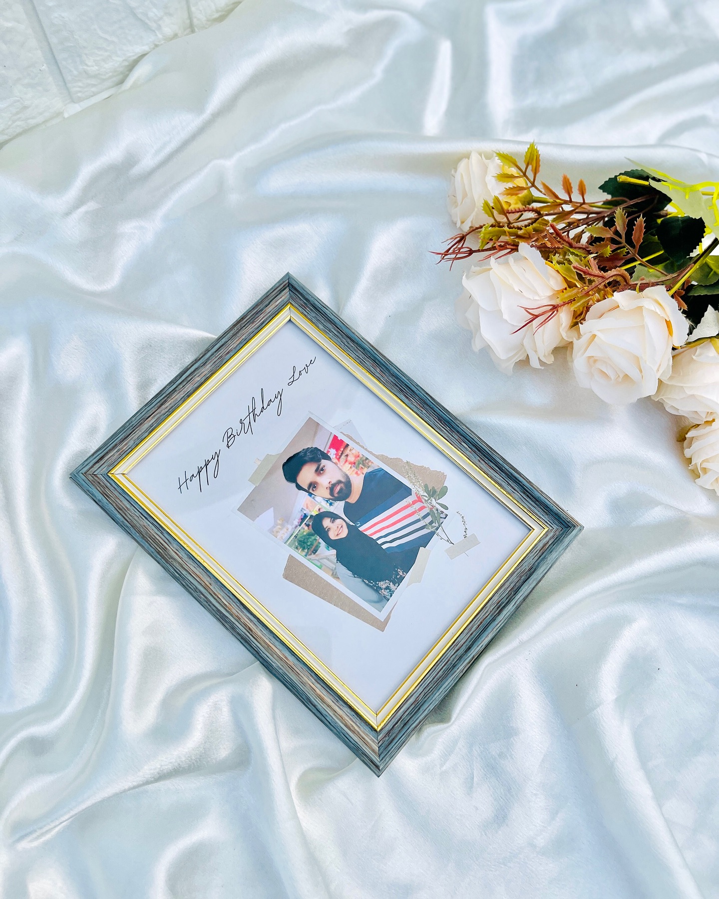 A wooden photo frame with a handwritten "Happy Birthday Love" message beside a soft bunch of white roses.