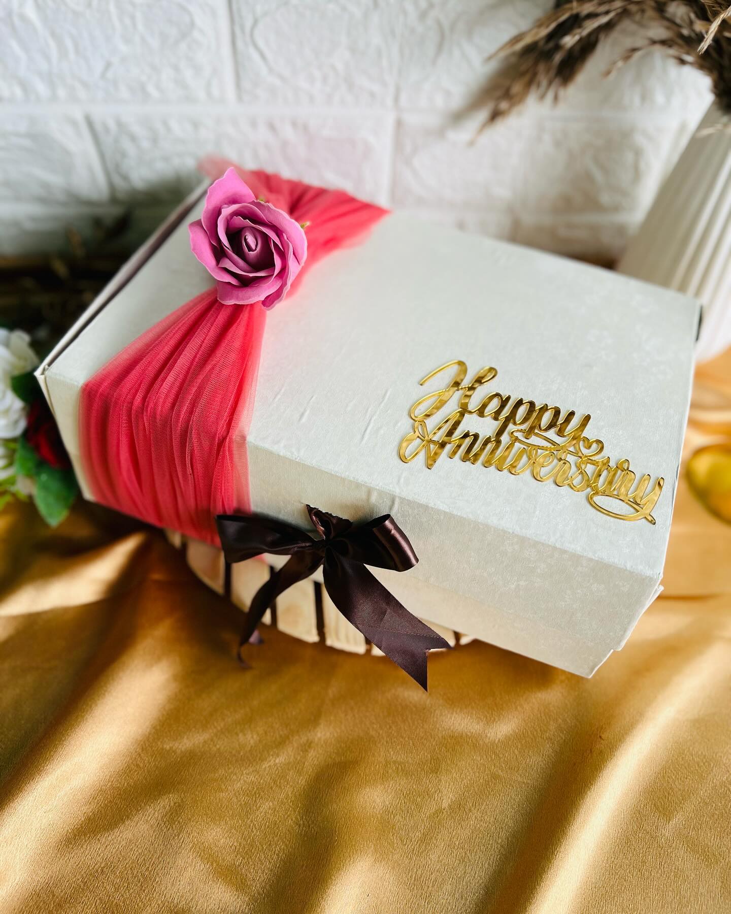 An ivory keepsake box wrapped in coral tulle with a blush rose and a gold Happy Anniversary topper.