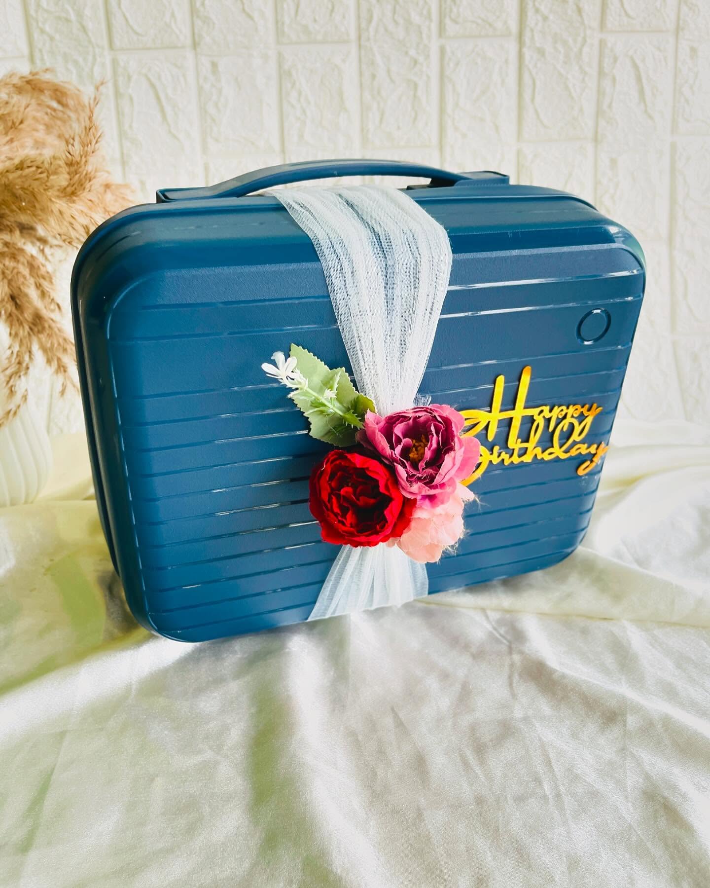 A teal suitcase-style gift box wrapped in soft tulle and red roses with a bright Happy Birthday topper.