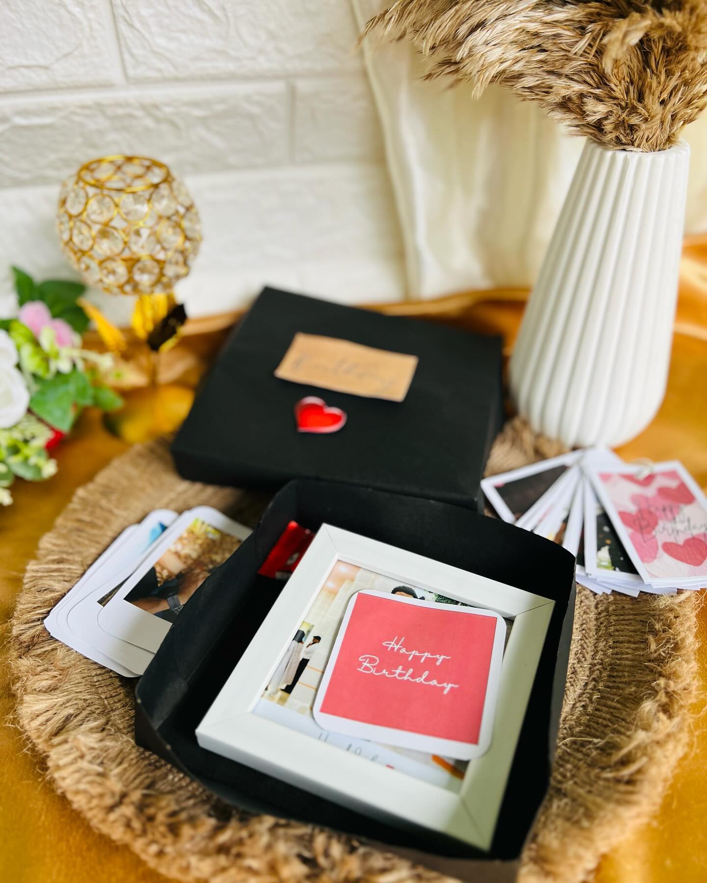 A rope-handled gift bag with a silk-bordered saree, a gold Happy Birthday topper, and a fresh pink rose.
