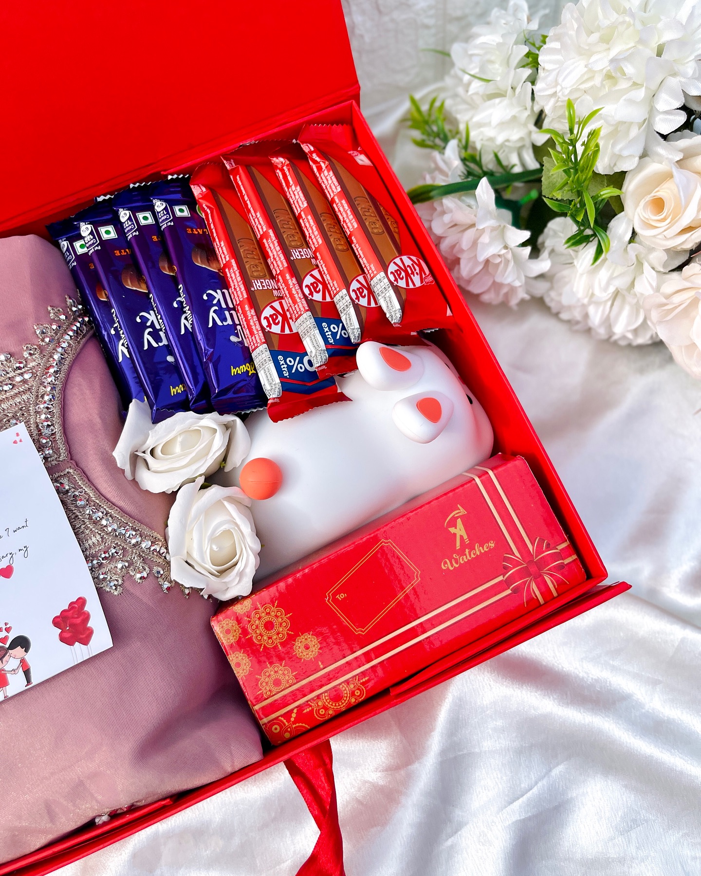 A red gift box pairing an embellished pink saree with Cadbury and KitKat chocolates, a soft toy, a watch box and a heartfelt anniversary card.