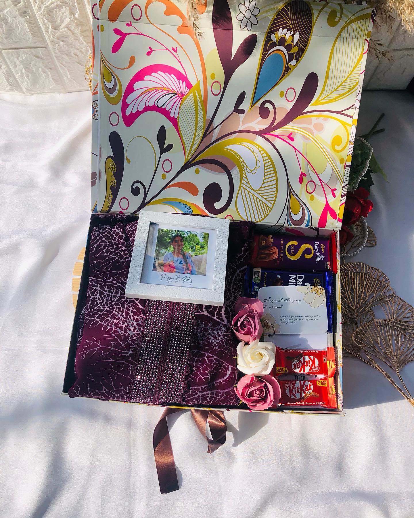 A patterned gift box with a maroon saree, assorted chocolates, fabric roses and a personalized photo frame.