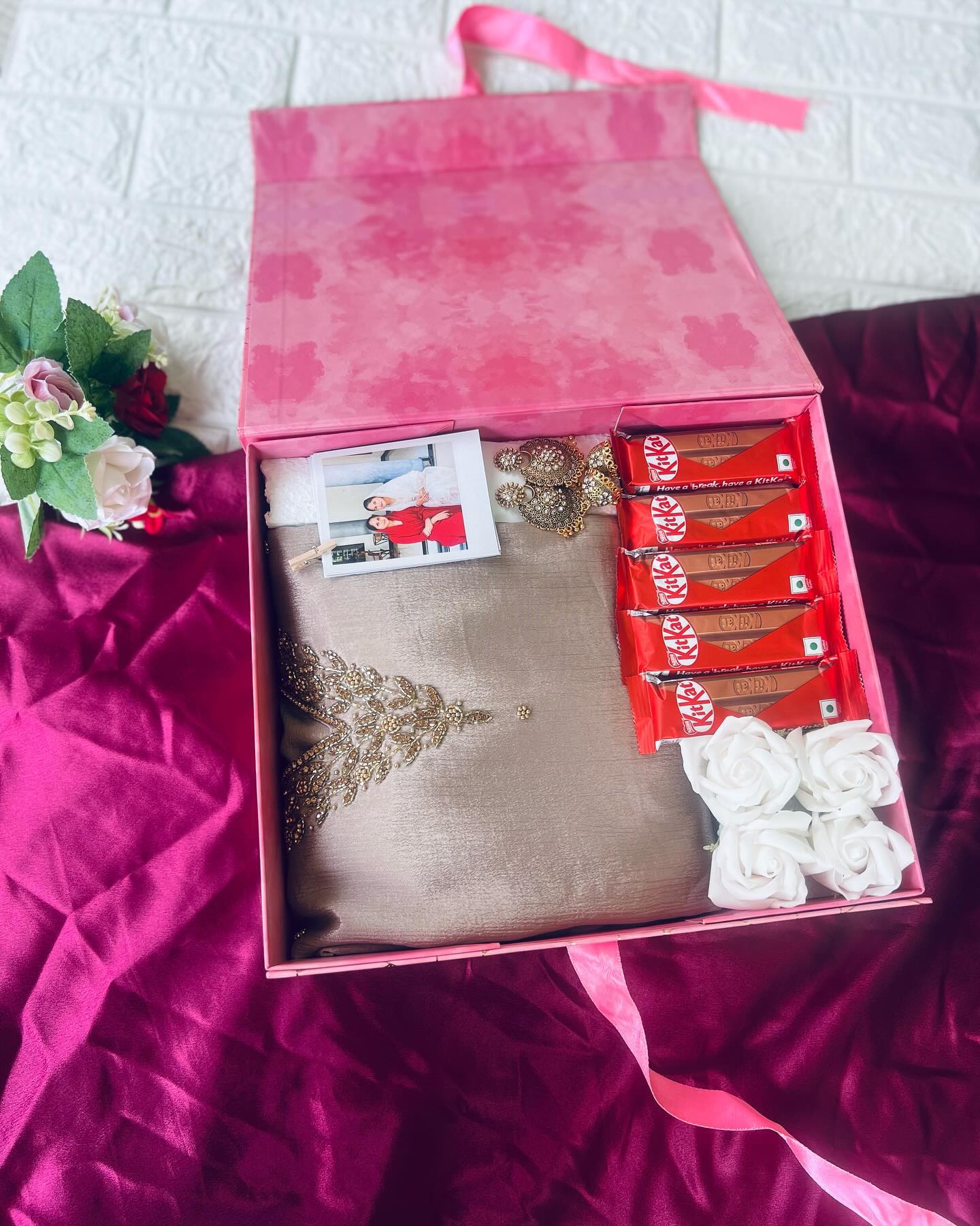 A pink gift box layered with an elegant saree, statement jewelry, KitKat bars, a photo, and soap roses.