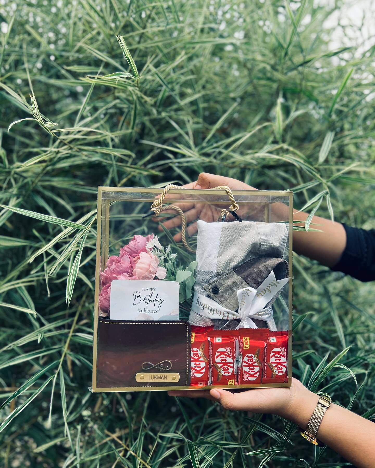 A clear birthday gift box featuring a monogrammed leather wallet, checkered shirt, fresh peonies, and KitKat bars.