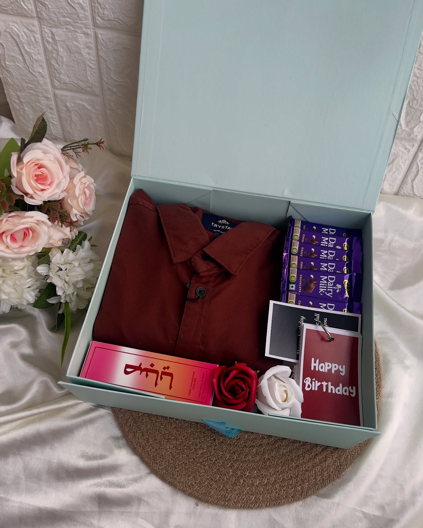 A birthday gift box for him with a maroon shirt, Dairy Milk chocolates, perfume, and roses.
