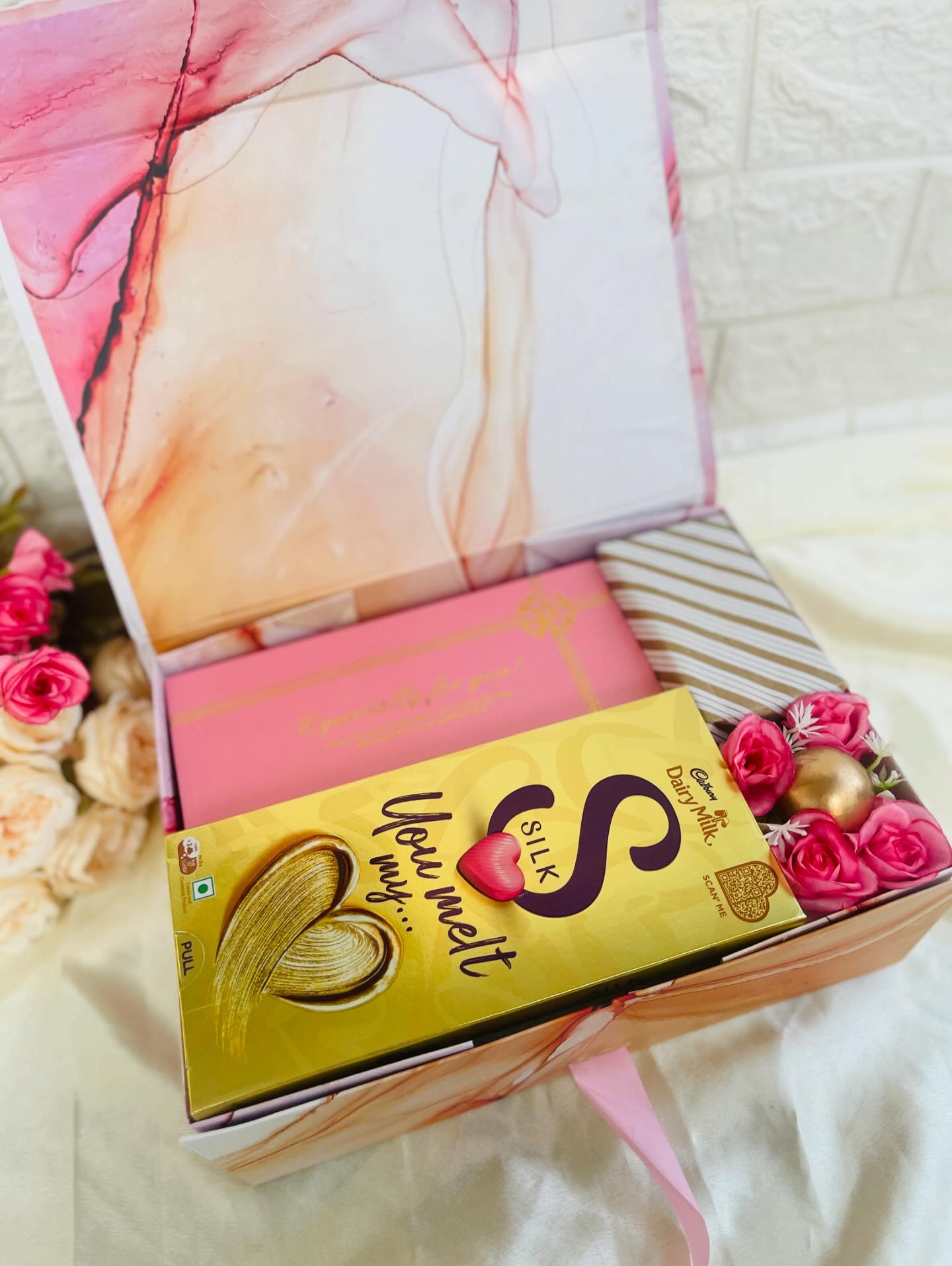 A marbled pink gift box filled with Cadbury Silk chocolate, a special-occasion treat box, and blush roses tied with gold ribbon.